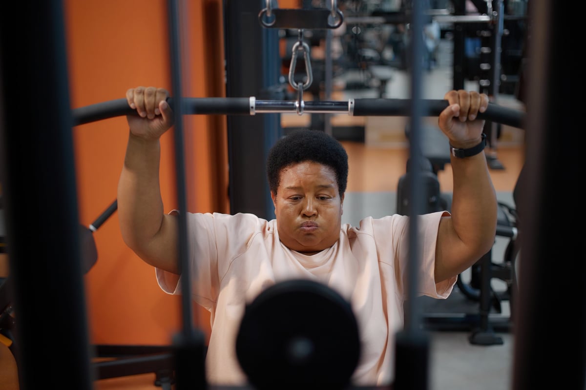 Portrait of middle aged Black woman exercising on strength training machine in gym, gripping handles and lifting weights, focused facial expression, fitness equipment visible in background