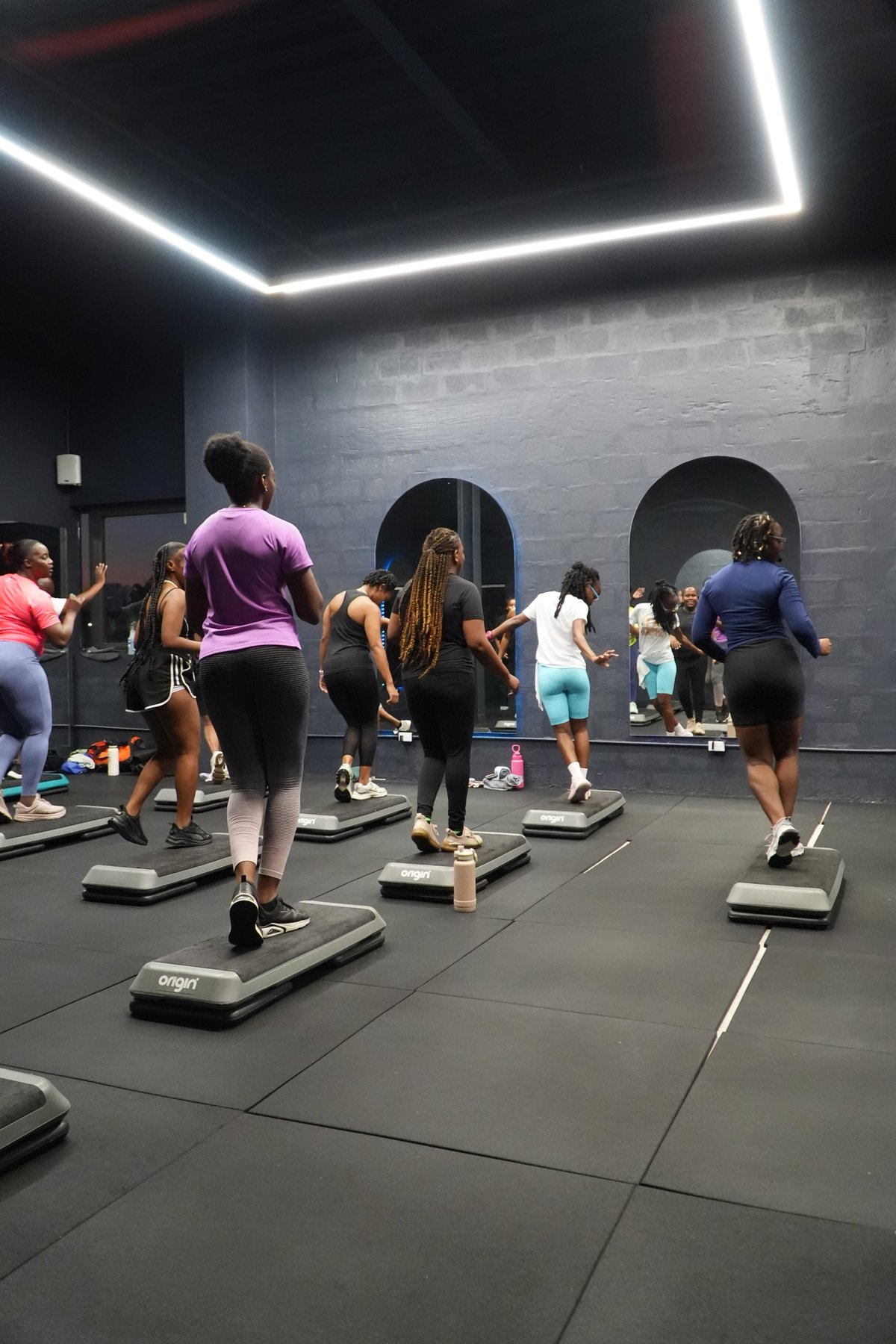 Group fitness class with participants on aerobic step platforms in a modern gym with neon ceiling lights and brick walls