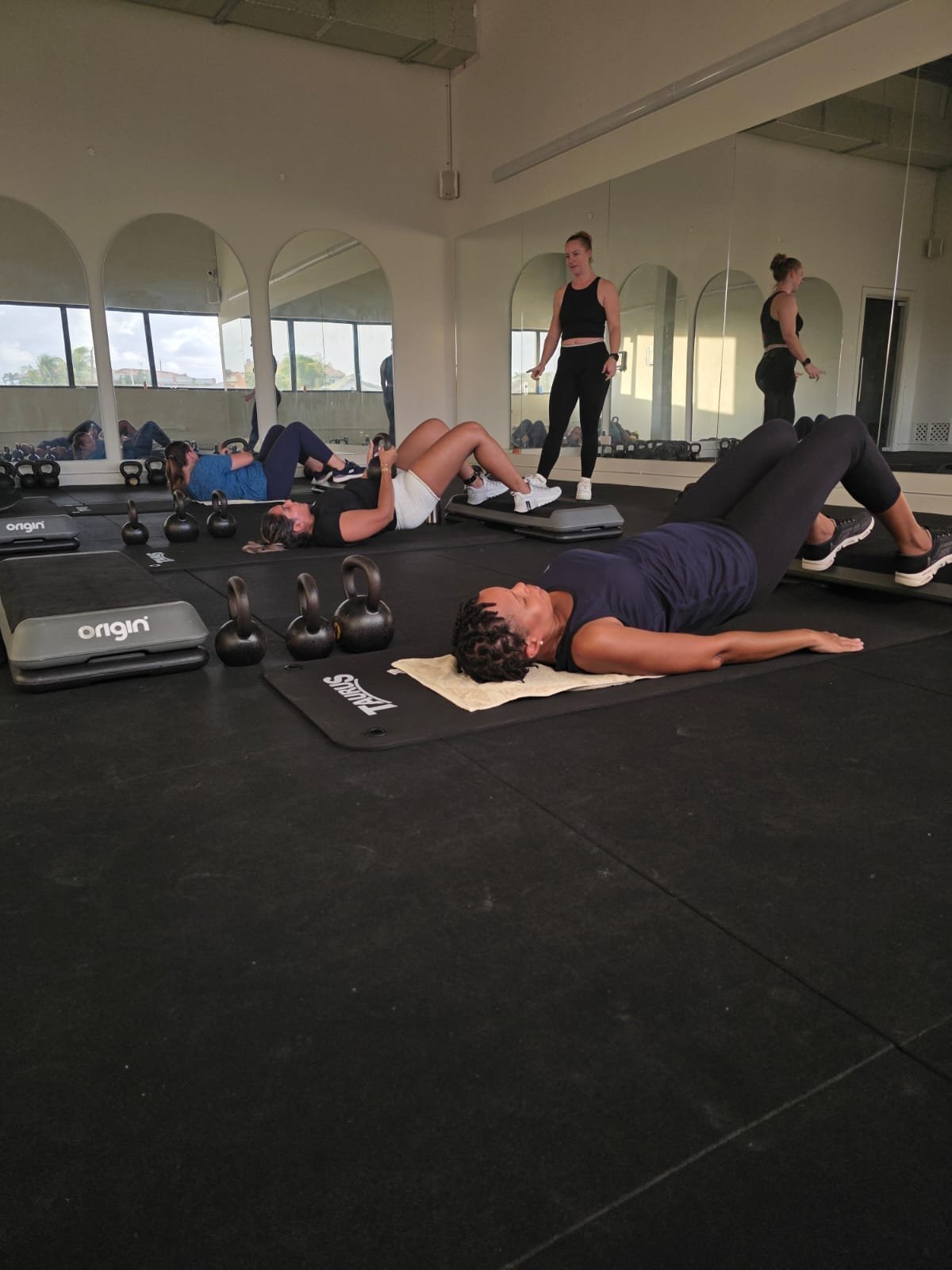 Group fitness class with people performing floor exercises on black mats in a modern gym studio with mirrored walls